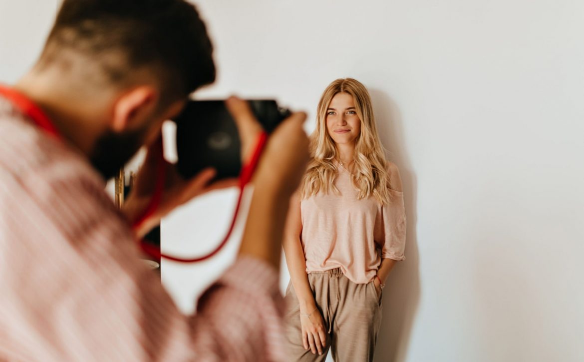 darkhaired-guy-makes-photo-his-blond-girlfriend-dressed-pink-tshirt-beige-pants-against-white-wall
